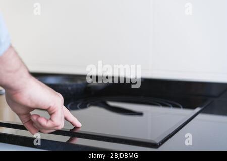 Mann, der in der Küche in einem Denim-Hemd kocht. Ein Anonymisierter drückt die Taste am Keramikkochfeld Stockfoto