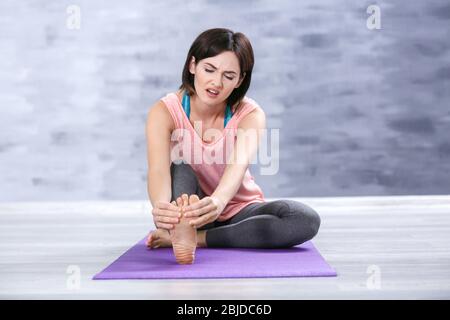 Schöne junge Frau, die Schmerzen im Fuß leidet, während sie drinnen auf Stretching-Matte sitzt Stockfoto