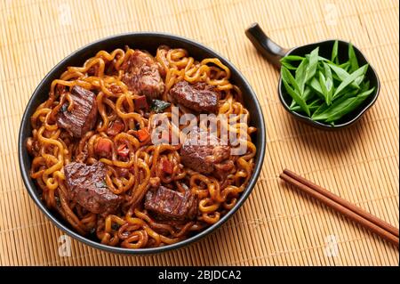 RAM-Don oder Chapaguri Nudeln mit Rindfleisch Steak in schwarzer Schüssel. Jjapaguri ist ein beliebtes südkoreanisches Gericht mit Ramen und udon Nudeln und Rindfleisch Steak. Stockfoto