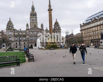 Zwei Männer, die durch einen menschenleeren George Square in Glasgow laufen, einer der beiden Männer trägt eine chirurgische Maske, um sich während der britischen Coronavirus Pandemie vor einer Infektion zu schützen. Stockfoto