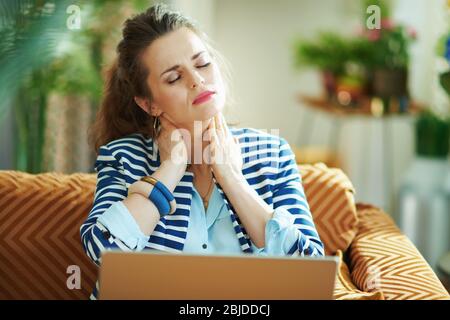 Betont stilvolle Frau in blauer Bluse und gestreiften Jacke mit Nackenschmerzen sitzen auf Couch mit Laptop bei modernen Hause in sonnigen Tag. Stockfoto