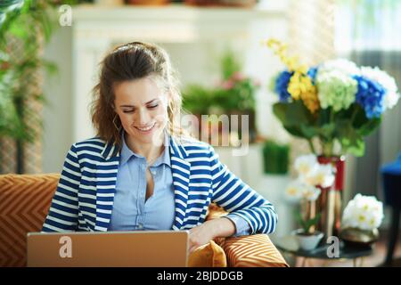 Lächelnde moderne 40 Jahre alte Frau in blauer Bluse und gestreiften Jacke sitzt auf Diwan mit Laptop im modernen Wohnzimmer bei schönem Tag. Stockfoto