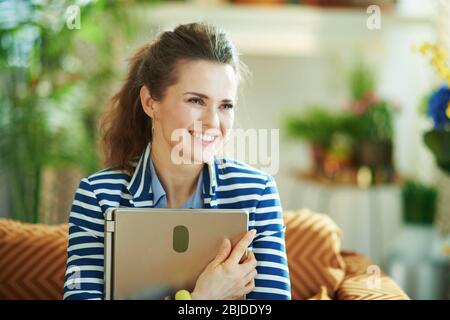 Lächelnd stilvolle Mittelalterhausfrau in blauer Bluse und gestreifte Jacke in der modernen Haus in sonnigen Tag umarmen Laptop. Stockfoto