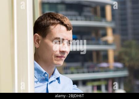 Ein 35-40-jähriger Kaukasischer steht auf dem Balkon und blickt auf das Stadtleben. Stockfoto