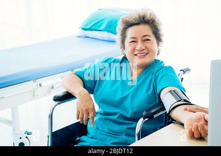 Eine lächelnde ältere weibliche Patientin, die auf einem Rollstuhl sitzt, wird von einer Krankenschwester oder einem Arzt im Krankenhaus oder in der Klinik Blutdruck gemessen Stockfoto