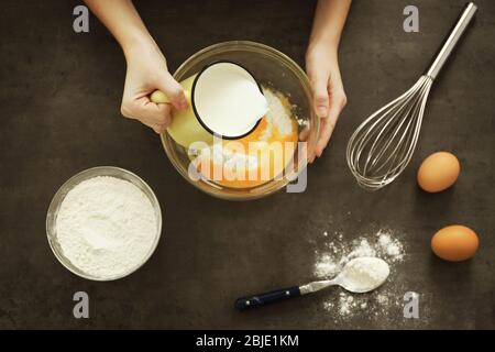 Weibliche Hände, die Milch in Glasschüssel mit Teig auf dem Tisch gießen, Draufsicht Stockfoto