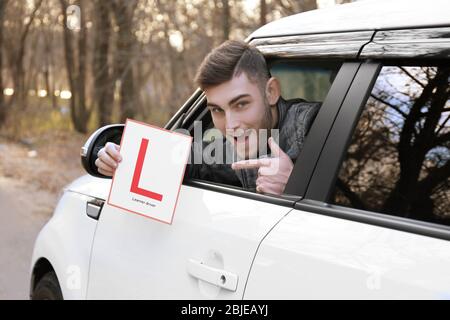 Junger Mann, der das Fahrerschild des Lernenden hält und aus dem Autofenster schaut Stockfoto