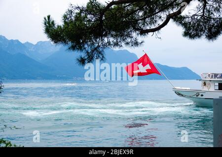 Schöne leuchtend rote Flagge der Schweiz winkt vor der Kulisse des Genfer Sees und Alpen, wie aus Villeneuve, Schweiz. Stockfoto