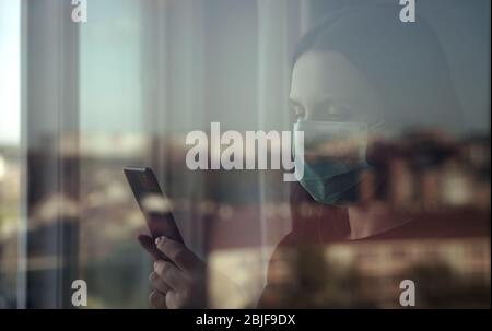 Frau mit Maske und Smartphone hinter geschlossenem Fenster Stockfoto