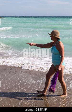 Eine Frau in einem hellblauen Badeanzug trägt ihre Muscheltasche und Punkte, während sie barfuß am Sandstrand von Fort Pierce, Florida, USA, entlang geht. Stockfoto