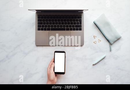 Top-Down-Ansicht einer Person, die ein Mobiltelefon über einem Schreibtisch mit einem leeren Bildschirm hält Stockfoto