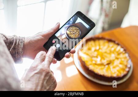 Hände einer alten Frau, die mit einem Smartphone zu Hause ein Foto von frisch gebackenem Apfelkuchen macht, um ihn in sozialen Netzwerken zu teilen. Konzept der alten Menschen und ich Stockfoto