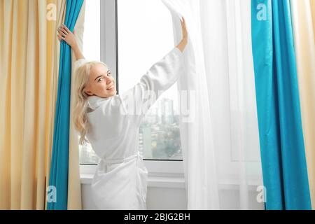 Junge Frau im Bademantel öffnen Vorhänge im Zimmer Stockfoto