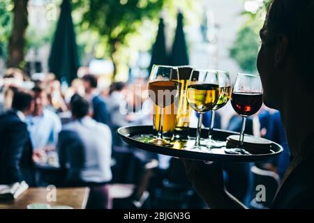 Nahaufnahme einer Frau, die ein Tablett mit Gläsern Rot- und Weißwein trägt. Bedienung im Cafe im Freien auf einem verschwommenen Hintergrund mit den Leuten. Stockfoto