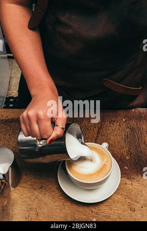 Barista macht Latte oder Cappuccino Kaffee, gießt gedämpfte Milch, macht Latte Art. Tasse Kaffee auf Holztisch. Stockfoto
