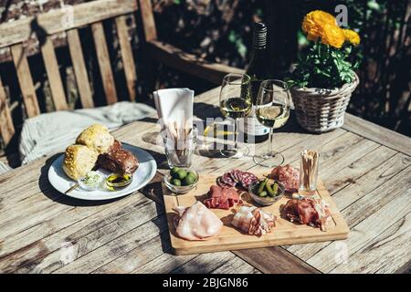 Charcuterie-Board mit Weißwein im Freien serviert. Eine Platte mit Schinken und Speck Scheiben mit Gurken auf einem Holzbrett Stockfoto