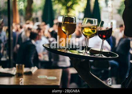 Nahaufnahme einer Frau, die ein Tablett mit Gläsern Rot- und Weißwein trägt. Bedienung im Cafe im Freien auf einem verschwommenen Hintergrund mit den Leuten. Stockfoto