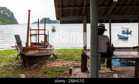 Frau in Sarong und weißen Fedora sitzt neben dem beacheten Fischerboot im Schutzraum Lesung mit traditionellen Fischerbooten im Hintergrund in Khao Lomm Stockfoto