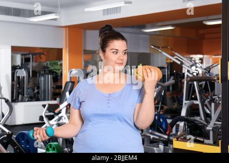 Übergewicht junge Frau essen Sandwich und halten Hantel im Fitnessstudio Stockfoto