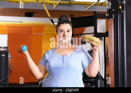 Übergewicht junge Frau essen Sandwich und halten Hantel im Fitnessstudio Stockfoto