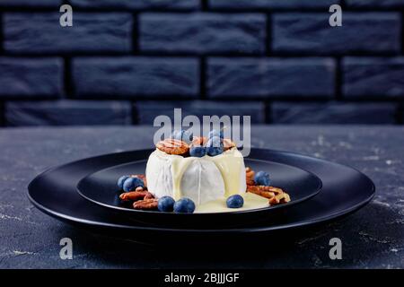 Französische klassische Küche gebratener Brie-Käse mit Nüssen und Beeren: Heidelbeeren, Preiselbeeren und Honig auf einem schwarzen Teller auf dem dunklen Beton serviert Stockfoto