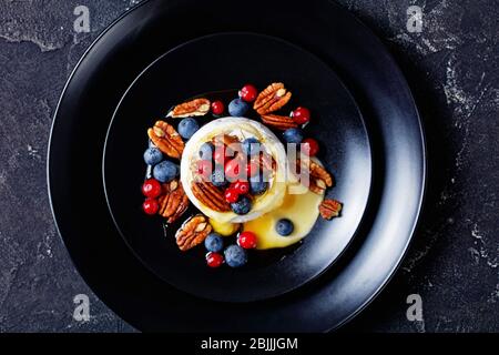 Runde Form ganze gebackene Brie mit Nüssen und Beeren: Heidelbeeren, Preiselbeeren und Honig auf einem schwarzen Teller auf einem dunklen Betongrund serviert, Stockfoto