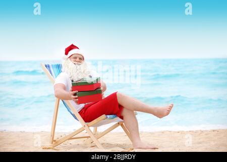 Authentischer Weihnachtsmann mit Geschenkboxen im Liegestuhl am Strand Stockfoto