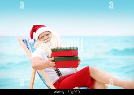 Authentischer Weihnachtsmann mit Geschenkboxen im Liegestuhl am Strand Stockfoto