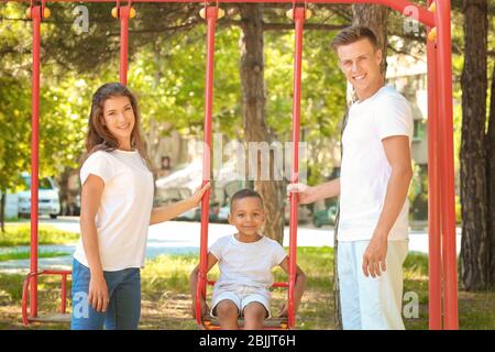 Junge Familie mit adoptierten afroamerikanischen Jungen spielen auf Schaukeln im Park Stockfoto