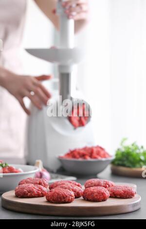 Koteletts aus Zangenfleisch auf Holzbrett und Frau mit Fleischwolf in der Küche Stockfoto