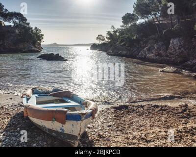 Verlassene Fischerboot in felsigen Bucht gegen Sonne. Felsige Wände mit privater Villa über der runden Bucht. Boot auf einem Sandstrand am Meer, Mallorca Insel Stockfoto