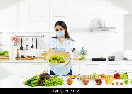 Junge Frau, die köstliche Speisen in der Küche kocht und Gesichtsschutzmaske trägt, um Speichel und Husten zu schützen. Bleiben Sie zu Hause während des Selbstquarans COVID-19 Stockfoto