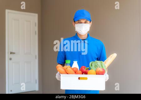 Asiatische Lieferung Mann trägt Gesichtsmaske und Handschuh mit Lebensmitteln Box mit Lebensmitteln, Obst, Gemüse und Getränk vor dem Kundenhaus während t stehen Stockfoto