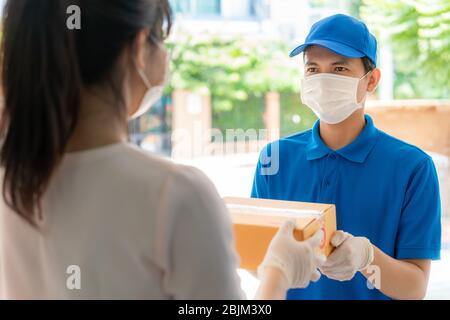 Asiatische Lieferung Mann trägt Gesichtsmaske und Handschuh in blauer Uniform hält einen Karton-Boxen im Vorderhaus und Frau eine Lieferung von Kisten von d Stockfoto