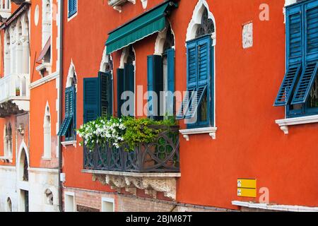 Auffällige und leuchtend orange gestrichene Außenwand mit schwarzen Holzfensterläden und weißen Zierleisten eines alten italienischen Hauses in Venedig, Italien. Balkon aus Metall überdacht Stockfoto