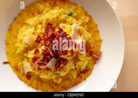 Safranrisotto aus nächster Nähe, klassisches gelbes buntes italienisches Grundgericht auf weißem Teller auf Holztisch mit Speck, ein Gourmet-raffiniertes Presenta Stockfoto