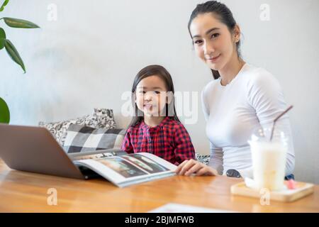 Asiatische Mütter lehren ihre Töchter, ein Buch zu lesen und Notebooks und Technologien für Online-Lernen während der Schulferien zu Hause zu verwenden. Educati Stockfoto
