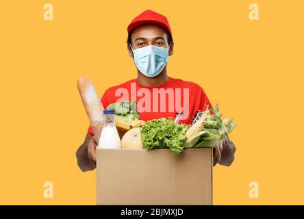 Delivery Concept - Handsome African American Delivery man mit Maske Tragepaket Box von Lebensmitteln und Getränken aus dem Geschäft. Isoliert auf gelb Stockfoto