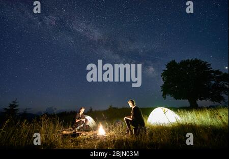 Wanderer sitzen vor einander auf einer Bank aus Baumstämmen und beobachten gemeinsam neben dem Lager und den Zelten in der Nacht das Feuer. Silhouette eines großen Baumes auf Hintergrund, Milchstraße und Sternenhimmel über ihnen Stockfoto