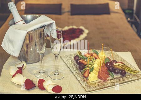 Hotel Suite für die Flitterwochen: Tisch mit Champagner und Obstteller, im Hintergrund ein Bett mit Rosenblättern verziert Stockfoto