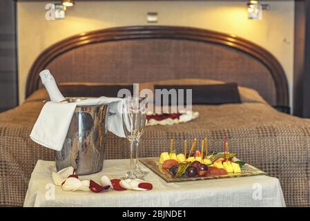 Hotelzimmer für die Flitterwochen: Tablett mit Champagner und Obstteller, im Hintergrund ein Bett mit Rosenblättern dekoriert Stockfoto