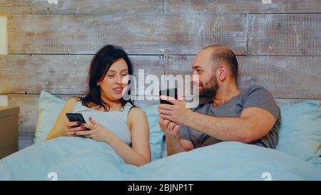 Fröhliches junges Paar mit Smartphone tragen Schlafanzug im Bett in der Nacht vor dem Schlafengehen. Stockfoto