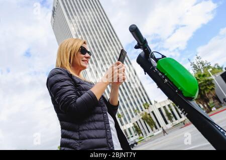 Schöne stilvolle Frau Scannen den qr-Code mit ihrem Telefon, um einen Elektroroller mit Wolkenkratzer Gebäude im Hintergrund mieten Stockfoto