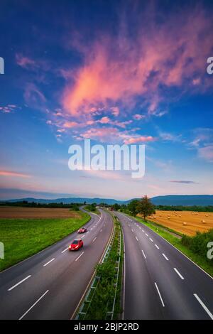 Breite, fast leere Straße unter dem bunten Himmel nach Sonnenuntergang, mit schönen roten Wolken und zwei roten Autos mit leichter Bewegungsunschärfe, ein dynamisches Transportat Stockfoto