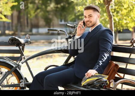 Junge gut aussehender Geschäftsmann Gespräch per Handy während sitzt auf der Bank im Park nach dem Reiten Fahrrad Stockfoto