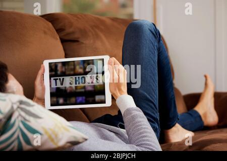 Nahaufnahme eines jungen kaukasischen Mannes, der bequem auf einer Couch liegt, sein Tablet anschaut, mit dem Text tv-Sendungen auf seinem Bildschirm geschrieben Stockfoto