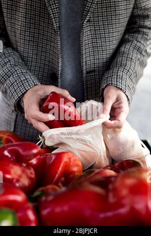 Nahaufnahme eines Mannes, der in einem Greengrocker einkauft und einige rote Paprika in einen wiederverwendbaren Textil-Netzbeutel stellt, um die Kunststoffverschmutzung zu verringern Stockfoto