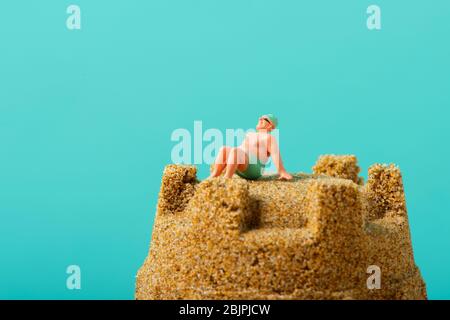 Nahaufnahme eines Miniaturmenschen in Badeanzug, der sich auf der Spitze einer Sandburg vor blauem Hintergrund mit etwas leerem Raum auf der Oberseite entspannt Stockfoto