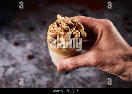 Nahaufnahme eines jungen kaukasischen Mannes mit einem Glas Dalgona-Kaffee in der Hand, vor einem grauen Hintergrund Stockfoto