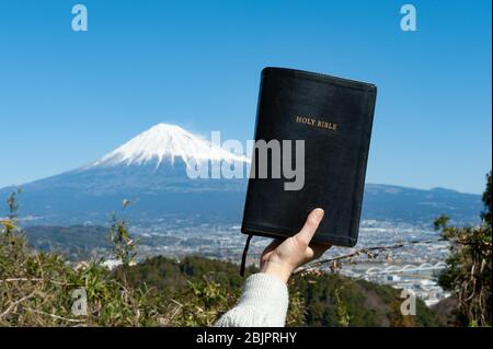 Erhobene Hand, die die Heilige Bibel hält. Hintergrund mit Luftaufnahme von Fuji City und Mount Fuji mit blauem Himmel an einem schönen Wintermorgen. Stockfoto